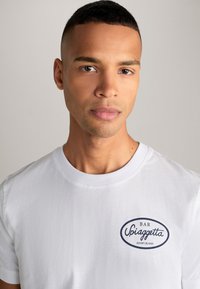 Weißes Baumwoll-T-Shirt mit Rundhalsausschnitt, mit einem dunkelblauen ovalen Logo auf der Brust, das "BAR Spiaggetta JOOP! JEANS" liest.