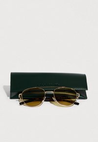 Okulary przeciwsłoneczne w kolorze złotym z brązowymi soczewkami na ciemnozielonym skórzanym etui z wytłoczonym napisem „Lacoste”. Okrągły kształt z akcentami w wzór żółwia.