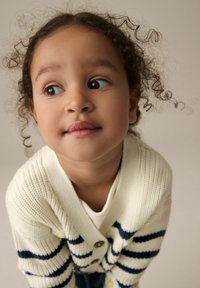 Hellbeige Strickjacke mit maritimen Streifen und großen Knöpfen. Das Kind hat lockige Haare und einen neutralen Gesichtsausdruck, vor einem sanften Hintergrund.