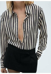 Gestreepte blouse in crème en marineblauw, met een diepe V-hals en knoopsluiting, gecombineerd met een hooggetailleerde zwarte rok. Glad textuur, relaxte pasvorm.