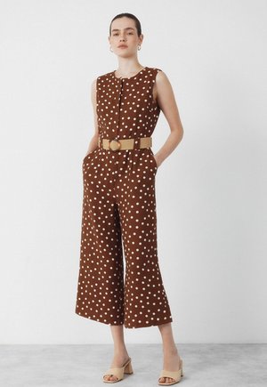 Kvinde iført brun ærmeløs jumpsuit med prikker, brede ben og tanfarvet bælte, stående med hænderne i lommerne og beige hælede sandaler.