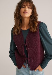 Gilet trapuntato bordeaux con motivo floreale, dotato di chiusura legata e colletto rotondo. Abbinato a una camicia a maniche lunghe teal e jeans in denim.