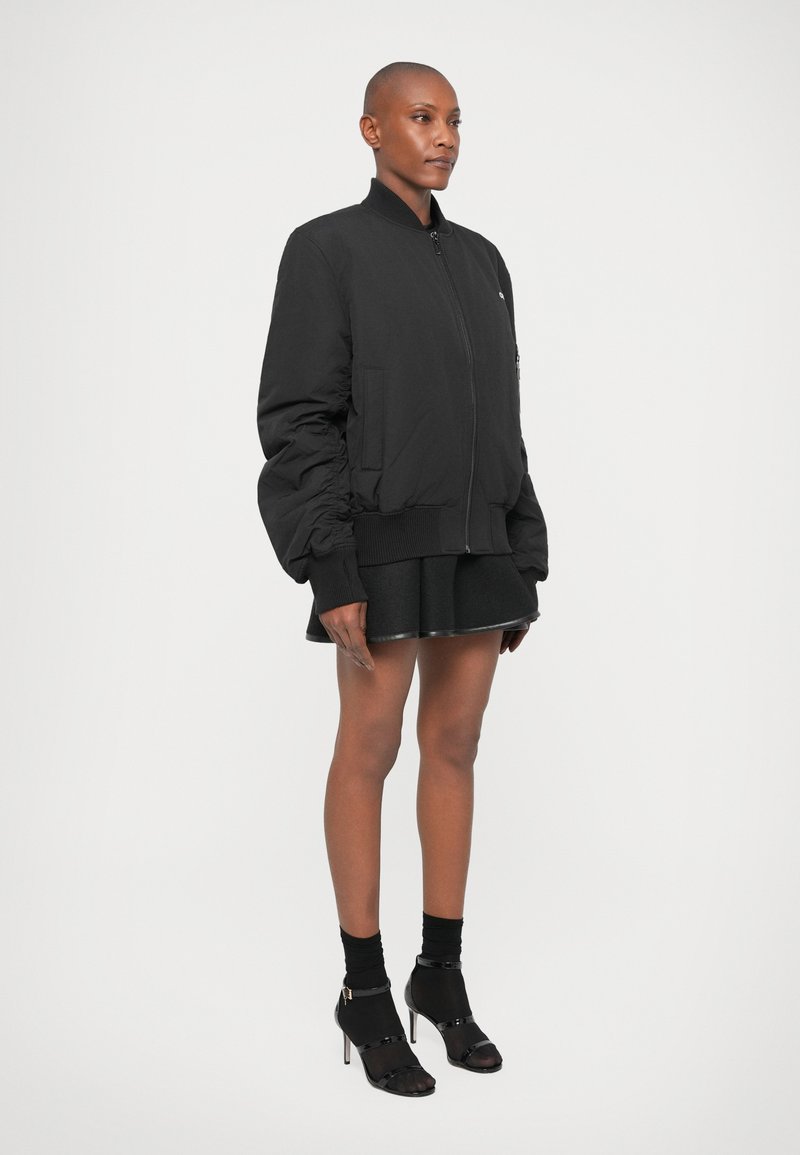 Veste bomber noire avec des poignets ajustés, fermeture éclair à l'avant et poches latérales. Associée à une jupe évasée noire et des chaussures à talons hauts et à bout ouvert.