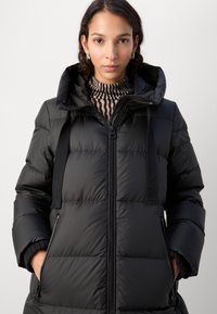 Schwarze Steppjacke mit hohem Kragen, fester Textur, Reißverschluss vorne und schmalen Trägern. Verfügt über seitliche Reißverschlusstaschen und ein gesteppetes Design.