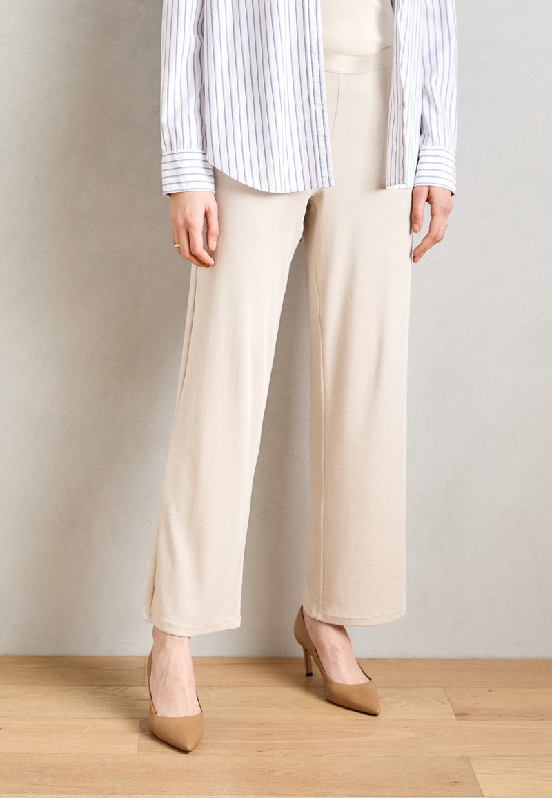 Personne portant un pantalon large beige, des escarpins à bout pointu couleur camel, et une chemise blanche à fines rayures verticales, debout sur un sol en bois.