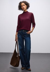 Un pull à col roulé bourgogne, un jean bleu foncé et un sac à épaule marron. Le pull a une texture lisse ; le jean est coupe droite, le sac est en daim.