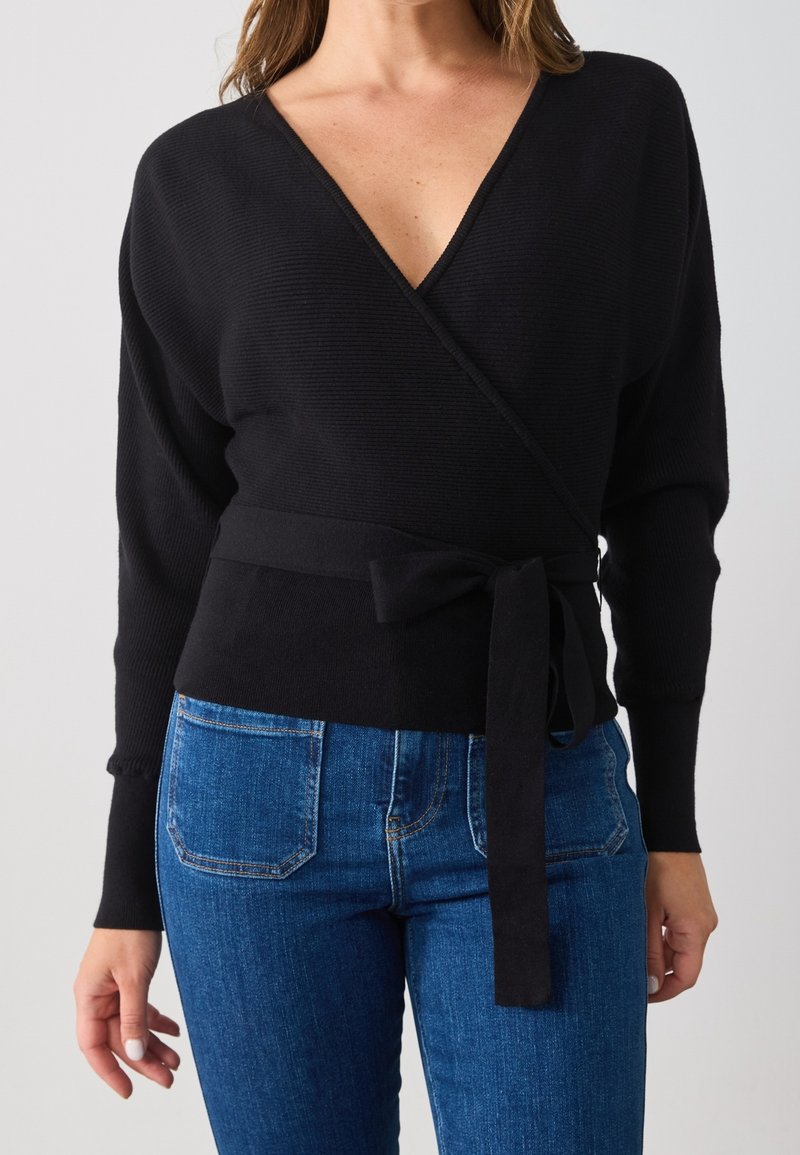 Schwarzer Wickelpullover mit tiefem V-Ausschnitt, langen Ärmeln und einer Schleife in der Taille. Gerippte Struktur kombiniert mit dunkelblauen Jeans.