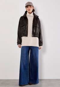 Braune Leder-Cropped-Jacke mit zwei Fronttaschen, übergroßer hellbeiger Strickpullover und weite blaue Jeans. Graue Karokappe.