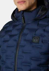 Navy Pufferjacke mit gestepptem Design, hohem Kragen, schwarzem Reißverschluss und einem Logo-Patch auf der Brust zur Markenkennzeichnung.