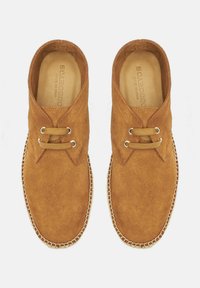 Zapatos de ante marrón con un diseño de corte bajo, dos ojales y un borde cosido. El interior está revestido con cuero beige.