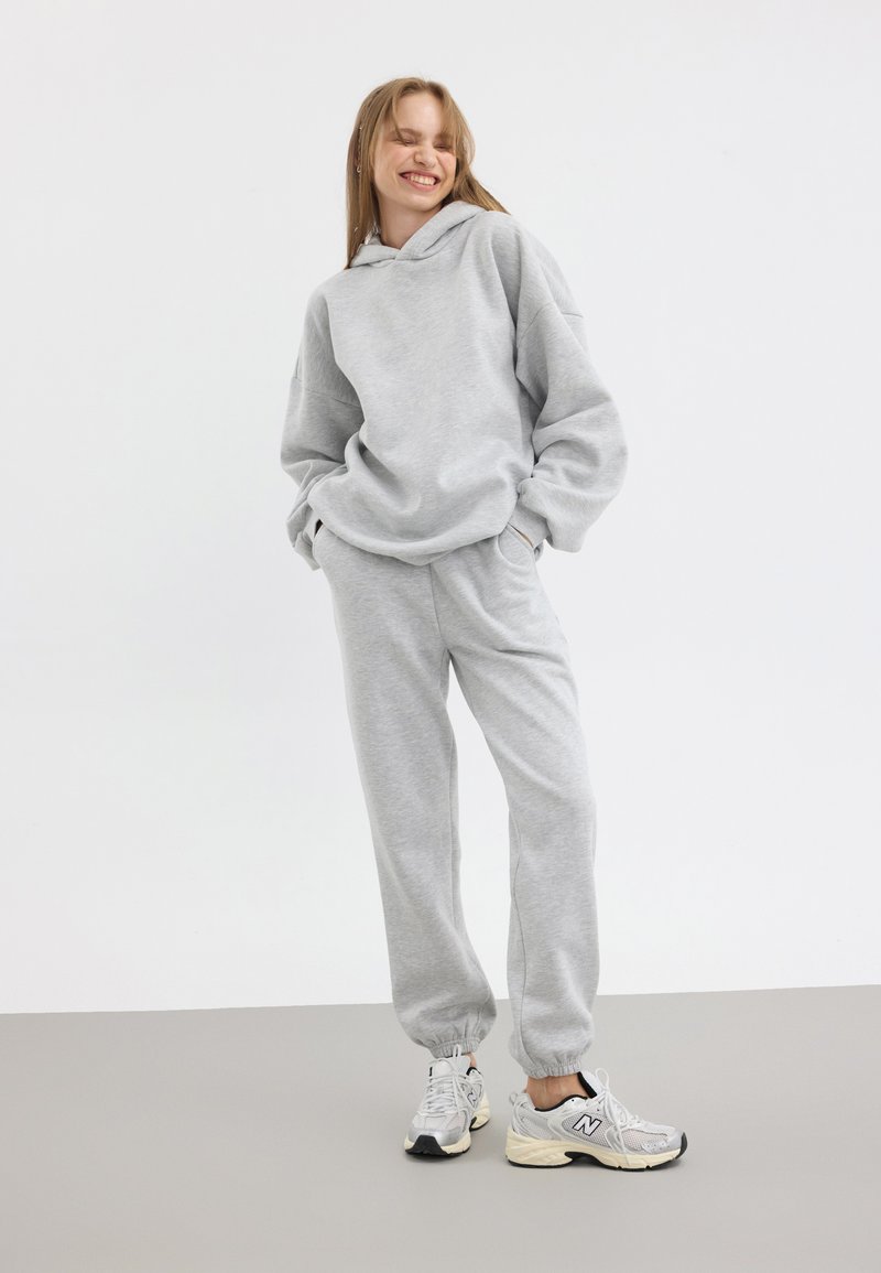 Grå hoodie med oversized passform och framficka, matchad med grå sweatpants. Modellen bär vita och grå sneakers.