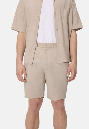 Hombre con pantalones cortos de lino beige y camisa de manga corta a juego abotonada sobre una camiseta blanca, frente a un fondo claro y liso.