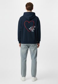 Felpa navy con grafica a cuore rosso e spille di sicurezza bianche sul retro. Tessuto morbido, vestibilità comoda, abbinata a jeans azzurri e sneakers bianche.