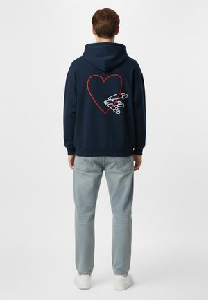 Hoodie bleu marine avec un graphique de cœur rouge et des épingles de sûreté blanches au dos. Tissu doux, coupe décontractée, associé à un jean bleu clair et des baskets blanches.