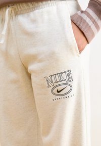 Világos bézs színű, puha anyagú melegítónadrág, rugalmas derékkal, fekete Nyomott Nike logóval és "SPORTSWEAR" felirattal.