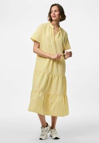 Vestido de algodón amarillo con cuello alto, mangas cortas y un diseño en capas. Cuenta con un detalle de cordón, combinado con zapatillas blancas.