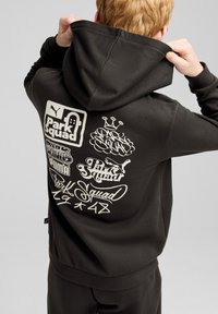 Sudadera negra con un gran estampado gráfico blanco que presenta "Park Squad" y varios logotipos en la parte posterior. El material parece suave y tiene un corte holgado.