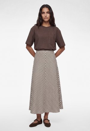 Woman wearing a brown pinstripe short-sleeve shirt, beige and brown chevron-patterned skirt, and brown Mary Jane shoes, standing with hands behind back.