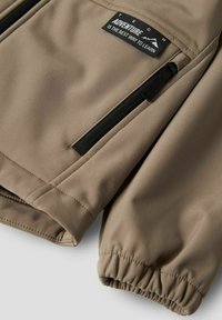 Gros plan sur la manche d'une veste beige avec un poignet élastique, une poche zippée noire et une étiquette portant l'inscription « L'aventure est la meilleure façon d'apprendre. »