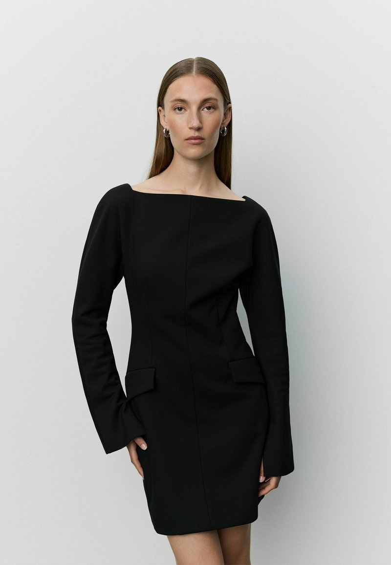 Femme aux longs cheveux lisses portant une robe noire cintrée à manches longues, avec un décolleté épaules dénudées et des poches avant à rabat, debout devant un fond uni.
