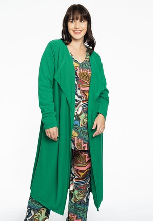 Cardigan vert avec un devant drapé et des manches longues, superposé à une tenue multicolore à motifs, comprenant des designs abstraits tourbillonnants.