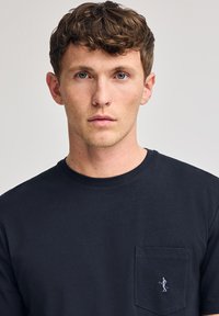 Camiseta de algodón azul marino con cuello redondo, mangas cortas y un bolsillo en el lado izquierdo del pecho que presenta un pequeño diseño bordado.