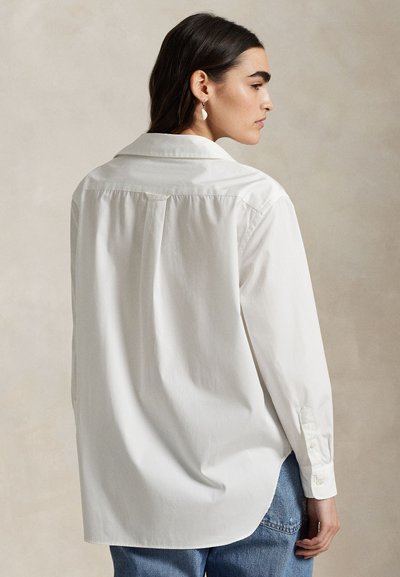 Naine tumedate juustega, kes kannab lahtist valget nööpidega särki ja siniseid teksaseid, näidatud tagant ja veidi küljele pööratud.