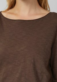 Zbliżenie osoby noszącej brązowy top z szerokim dekoltem, ukazujące linię szyi, obojczyk i część długich blond włosów.