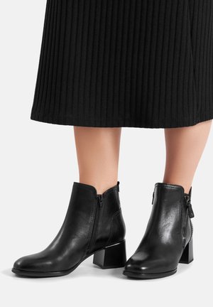 Czarne skórzane botki na słupku, z zapięciem na zamek błyskawiczny i gładką fakturą, prezentowane obok plisowanej czarnej spódnicy.