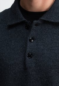 Dunkelgraues Wollpolohemd mit Kragen, drei schwarzen Knöpfen und strukturiertem Stoff. Nahaufnahme des Halsbereichs.