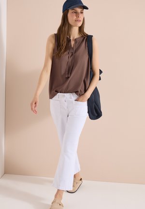 Femme marchant portant un haut marron sans manches, un pantalon blanc, des chaussures beige à enfiler, une casquette bleu marine et un sac bleu marine porté sur une épaule.