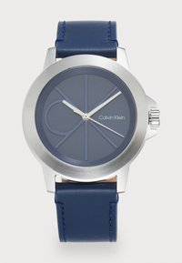 Montre Calvin Klein avec un boîtier rond en acier inoxydable, un bracelet en silicone bleu marine, un cadran bleu avec des marquages géométriques et des aiguilles argentées.