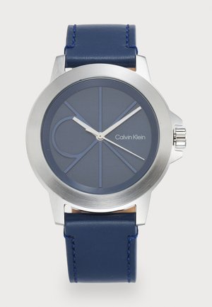 Montre Calvin Klein avec un boîtier rond en acier inoxydable, un bracelet en silicone bleu marine, un cadran bleu avec des marquages géométriques et des aiguilles argentées.