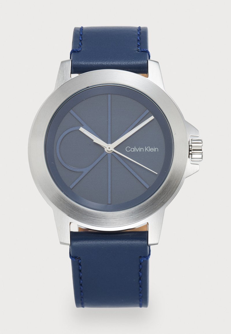 Zegarek Calvin Klein z okrągłą, stalową kopertą, granatowym silikonowym paskiem, niebieską tarczą z geometrycznymi oznaczeniami i srebrnymi wskazówkami.