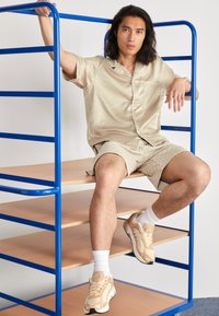 Chemise beige clair à manches courtes et shorts assortis avec un motif subtil. Porte des baskets crème et des chaussettes blanches de type crew, assis sur une étagère en métal bleu.