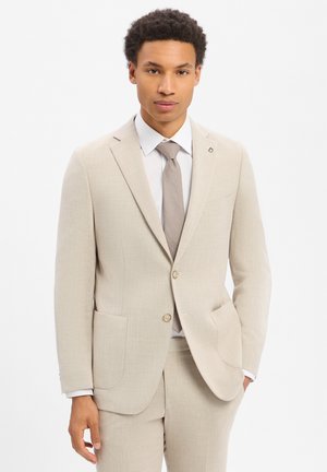 Mann trägt einen beigen Anzug, weißes Hemd und gemusterten beigen Krawatte, steht mit einer Hand in der Tasche vor einem schlichten weißen Hintergrund.