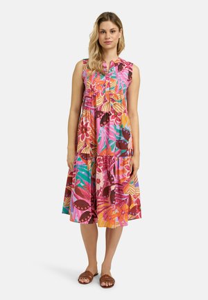 Woman wearing a sleeveless, multicolored floral midi dress with a tiered skirt and brown flat sandals, standing against a plain background.