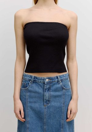 Kobieta w czarnym prążkowanym, bezramkowym topie odsłaniającym brzuch, w połączeniu z niebieską spódnicą z wysokim stanem, stojąca na jednolitym tle.