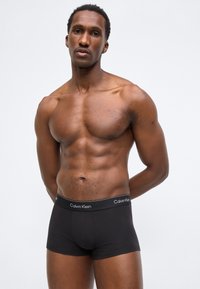 Μυώδης άνδρας που στέκεται με τα χέρια πίσω από την πλάτη, φοράει μαύρα μπόξερ Calvin Klein, σε απλό λευκό φόντο.