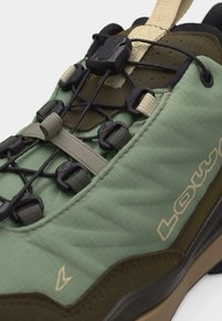 Green and brown athletic shoe with quick-lace system featuring black adjustable toggles and fabric pull tab on tongue.