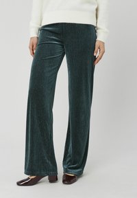 Pantalon large en velours bleu sarcelle avec nervures verticales, offrant une texture douce et un éclat subtil. Associé à des chaussures vernies foncées.