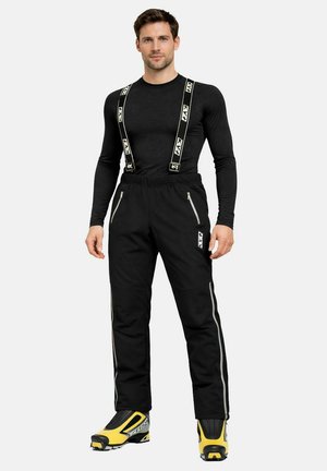 Mann trägt ein schwarzes Langarmshirt, schwarze Skihosen mit Hosenträgern und gelb-schwarze Skischuhe, steht vor weißem Hintergrund.