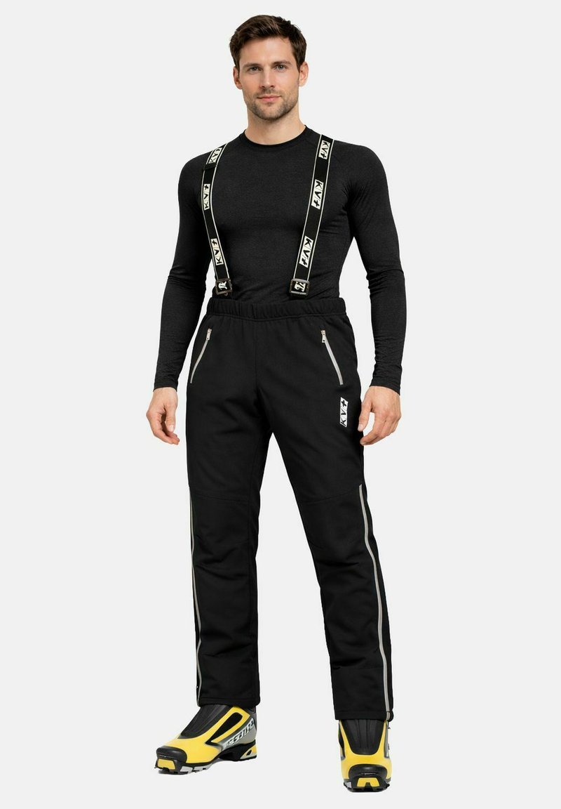 Homme portant une chemise noire à manches longues, un pantalon de ski noir avec des bretelles, et des chaussures de ski jaunes et noires, debout sur un fond blanc.