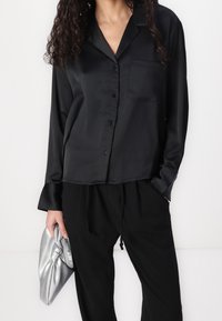 Svart satin skjorta med knappar, långärmad, bröstficka och slät yta. Kombineras med svarta löst sittande byxor och en silverfärgad clutch.