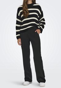 Maglione a righe bianche e nere in maglia con maniche lunghe, abbinato a pantaloni neri e sneaker bianche su un modello.