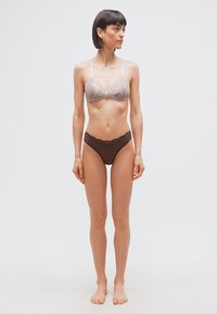 Γυναίκα με κοντά σκούρα μαλλιά φορώντας μπεζ δαντελένιο σουτιέν και σκούρο καφέ εσώρουχο με δαντέλα, όρθια ξυπόλητη μπροστά σε απλό λευκό φόντο.