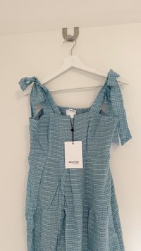 Kék kockás ruha vállpántokkal, texturált anyagból, fodros részletekkel és a dekoltázsról lógó címkével, akasztón.
