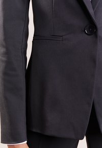 Schwarze Sakkokjacke mit glatter Textur, ein Knopfverschluss und taillierte Passform. Verfügt über eine Seitentasche und einen subtilen Glanz.