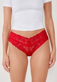 Perizoma rosso in pizzo con motivi floreali, caratterizzato da un design a vita alta e bordi scallopati, che evidenzia una trama e dettagli intricati.