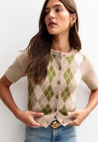 Una donna con lunghi capelli castani indossa un cardigan beige e verde a rombi con bottoni, jeans blu e orecchini a cerchio dorati, mentre sistema il suo maglione.